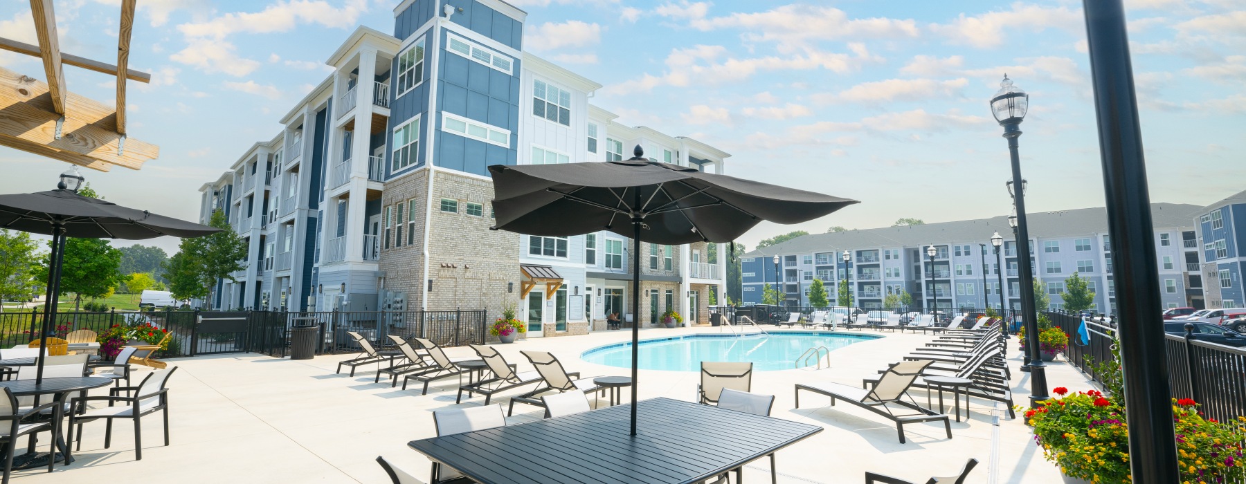 An outdoor pool area with lounge chairs, umbrellas, and patio tables in front of modern apartment buildings under a partly cloudy sky.
